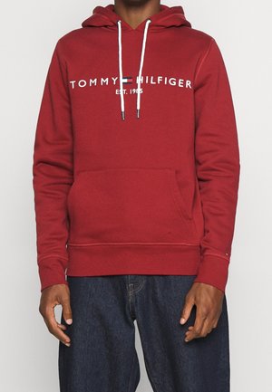 Homme portant un hoodie rouge Tommy Hilfiger avec des cordons blancs et un jean bleu, se tenant contre un fond clair.