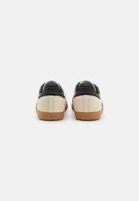 Sneakers with a cream upper, black accents, and a textured brown rubber sole, featuring stitched detailing on the heel and tongue.