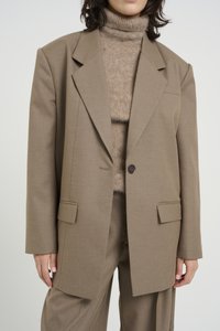 Beige oversized blazeri teksturoiduss kangas, millel on soonilised kraed, ühe nööbi kinnitamine ja küljetaskud, kantud heledast pruunist polo-särgi peal.