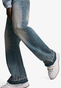 Hellblaue Denim-Jeans mit entspannter Passform, ausgefranstem Saum und subtilen Verblassungen. Kombiniert mit weißen Turnschuhen, die einen lässigen Stil zeigen.