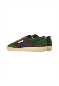 Zapatillas de ante verde con acentos marrones y marinos, detalles perforados, suela plana beige y una etiqueta lateral con el nombre de la marca.