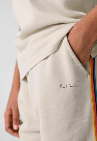 Close-up af en hånd i lommen på beige joggingbukser med flerfarvet stribet kant og "Paul Smith" logo broderet på låret.