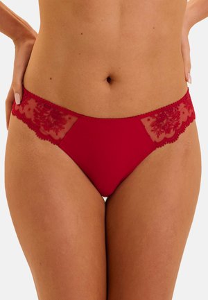 Woman wearing red lace bikini-style underwear with floral embroidery and semi-sheer side panels, hands resting on hips.