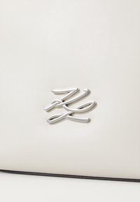 Bílá kožená taška se stříbrným metalickým logem na přední straně, s hladkou texturou a minimalistickým designem.