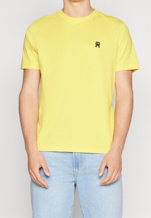 Yellow cotton t-shirt with a round neckline and short sleeves. Features a small black logo on the chest. Worn with light blue jeans.