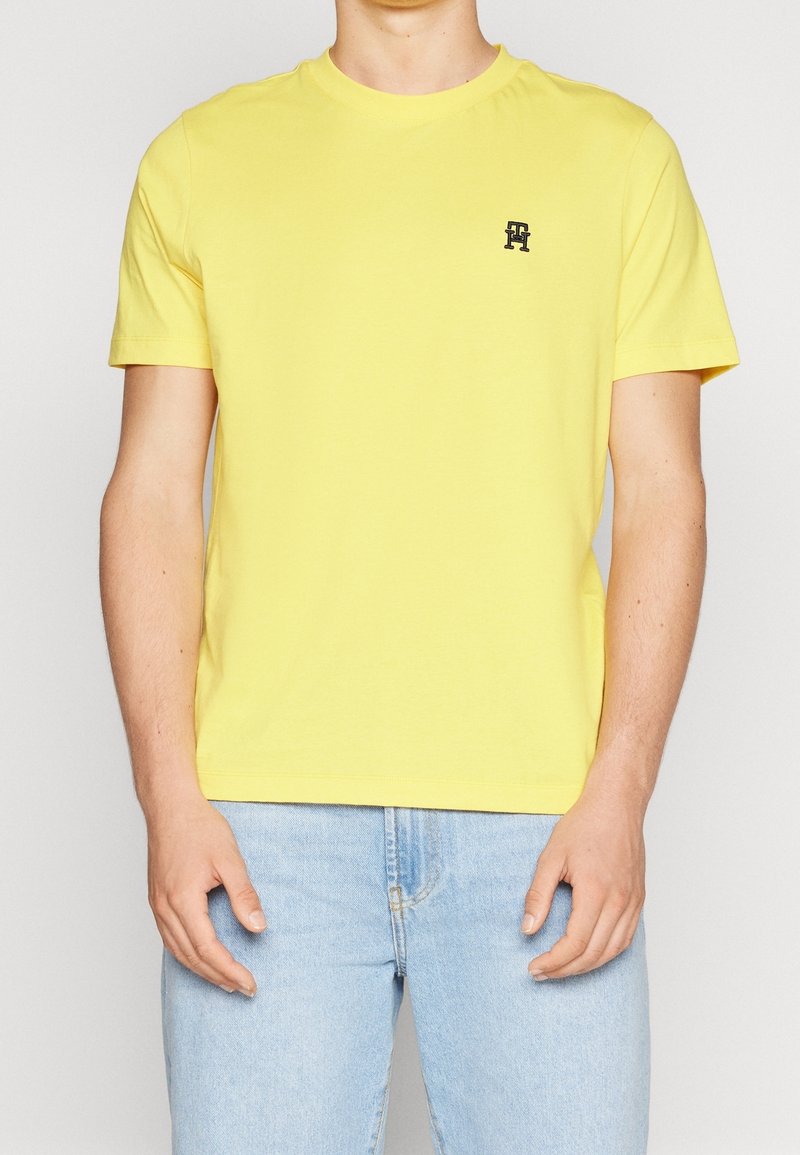 Yellow cotton t-shirt with a round neckline and short sleeves. Features a small black logo on the chest. Worn with light blue jeans.
