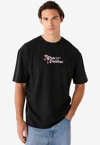Must musta t-särk, millel on roosa graafiline trükk ja tekst "Pink Panther", lühikesed varrukad ja mugav lõige. Pole eristuvaid mustreid ega aktsente.