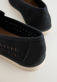 Zapatos de ante negros tipo slip-on con detalles tejidos a los lados, suelas de goma color crema y costuras azules en contraste alrededor de los bordes.