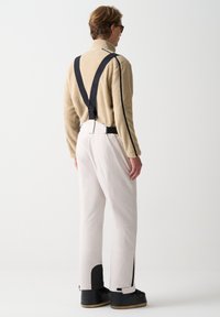 Béžový fleecový pulovr s vysokým límcem; bílé lyžařské kalhoty se zipy na bocích; černé šle s logem; černé boty s kovovými detaily.