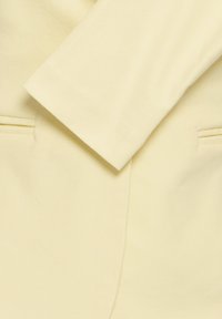 Tissu de couleur crème avec une manche de vêtement pliée et des détails de couture, montrant une partie de deux poches passepoilées horizontales.