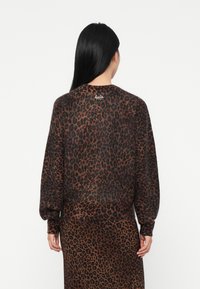 Mulher com cabelo longo e negro vestindo um suéter de padrão leopardo castanho e preto e uma saia de leopardo a condizer, de costas sobre um fundo branco.