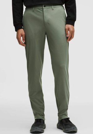 Egy férfi olíva zöld slim fit nadrágban és fekete tornacipőben, kezét lazán oldalán tartva áll.