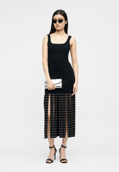 Γυναίκα με μαύρο φόρεμα χωρίς μανίκια και φούστα με μοτίβο και σχίσιμο, μαύρα πέδιλα με ψηλό τακούνι, μαύρα γυαλιά ηλίου και ασημί τσαντάκι χειρός.