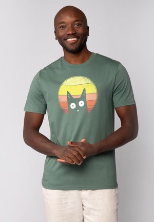 Groene katoenen t-shirt met een afbeelding van een zwarte kat met grote ogen, tegen een ronde gele en roze zonsondergang achtergrond.