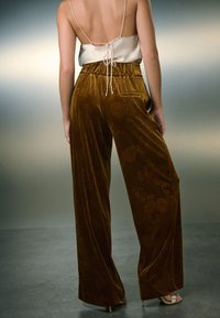 Pantalones anchos de terciopelo marrón con textura floral, cintura elástica y detalle de bolsillo trasero. Un diseño de silueta holgada completa el conjunto.