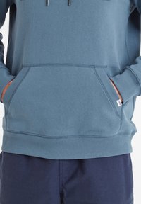 Blue cotton sweatshirt with a front pocket, ribbed cuffs, and hem. Silver drawstrings accent the hood. Worn with dark navy shorts.