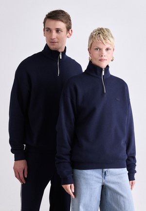 Navy blue zip-up sweatshirt made of soft fabric with a high collar. Features long sleeves and a logo on the chest. Paired with light jeans.