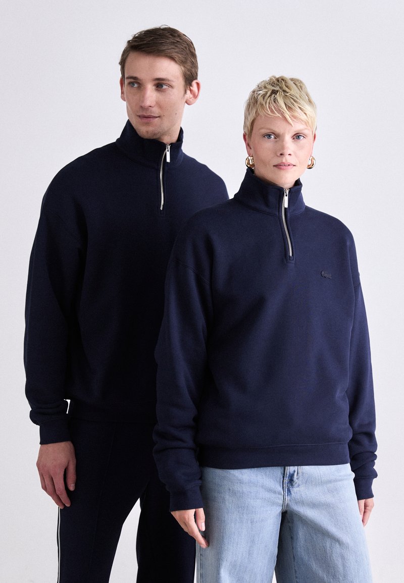 Tamsiai mėlynas zip-up džemperis, pagamintas iš minkštos medžiagos su aukštu apykakle. Turi ilgas rankoves ir logotipą ant krūtinės. Derinamas su šviesiomis džinsomis.