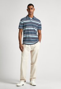 Svītraina polo krekls zilā, baltā un jūras spēku krāsā, ar apkakli. Tas valkāts kopā ar gaiši pelēkiem biksēm un baltām sporta apaviem uz neitrāla fona.