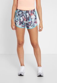 Person som bär flerfärgade sportshorts med blå kant, en persikofärgad ärmlös topp och vita löparskor, står på ett vitt golv.