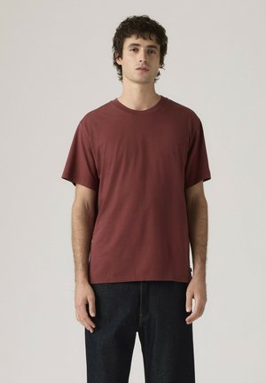 Junger Mann steht, trägt ein einfaches bordeauxrotes kurzärmeliges T-Shirt und dunkle blaue Jeans vor einem hellgrauen Hintergrund.