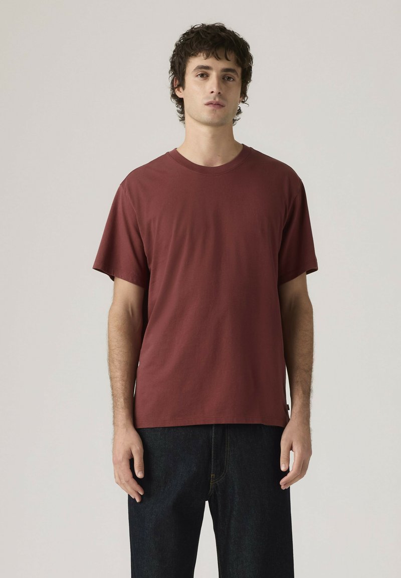 Giovane in piedi, indossa una maglietta a maniche corte color bordeaux semplice e jeans blu scuro su uno sfondo grigio chiaro.