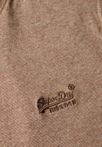 Tela a rayas marrón con una superficie texturizada. Logo bordado de "Superdry" en marrón más oscuro en la esquina inferior derecha.