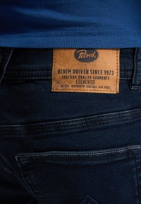 Jeans de mezclilla con una tela azul oscura, que presentan un parche de cuero marrón con la marca "Petrol" y texto sobre la calidad de la artesanía.
