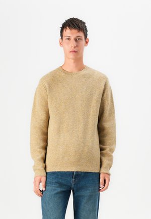 Beige ribbad stickad tröja med rund halsringning och långa ärmar, kombinerad med blåjeans. Mjuk textur och avslappnad design.