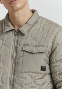Veste matelassée marron clair avec fermeture éclair, col montant et une poche à rabat avec une étiquette de marque sur le côté gauche. Tissu lisse et texturé.