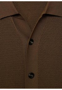 Polo de punto marrón texturizado con cuello, presenta un cierre de botones negros y un patrón sutil en toda la tela.