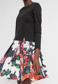 Svart långärmad topp med rund halsringning, parat med en veckad blommig kjol med röda, gröna och svarta mönster på vit bakgrund.