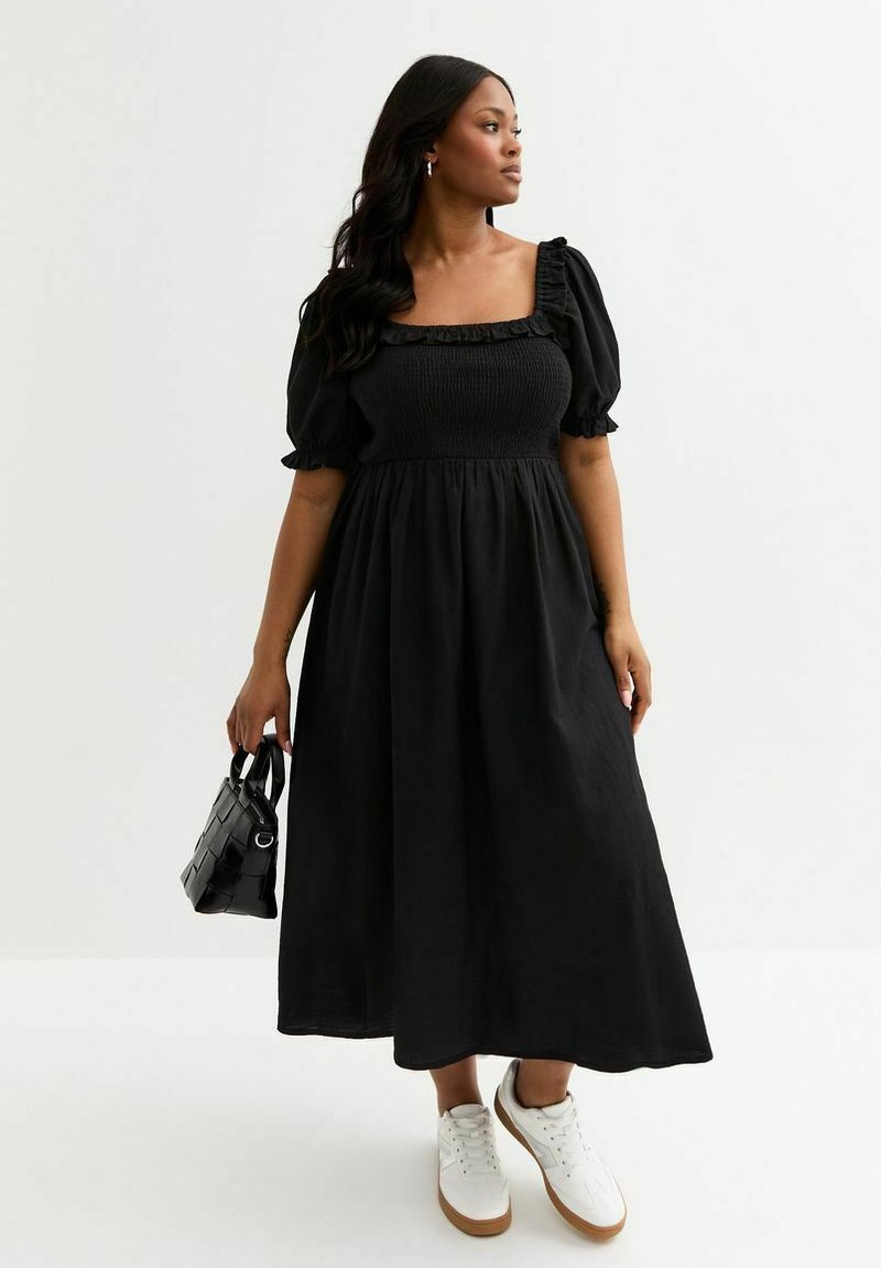 Vestido negro con escote cuadrado, mangas abullonadas y cuerpo fruncido, que llega hasta la mitad de la pantorrilla. Combinado con zapatillas blancas y un pequeño bolso negro.