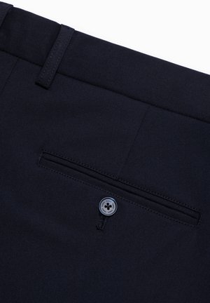 Granatowe spodnie z płaskim przodem, z tylną kieszenią zapinaną na guzik i gładką teksturą materiału. Ujęcie z góry.