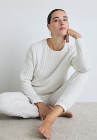 Ung kvinna sittande på en matta, iklädd vit vadderad loungekläder, lutar huvudet mot handen, med neutral uttryck och bakåtkammande hår.