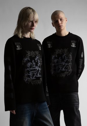 Doi tineri adulți purtând hanorace negre cu imprimeuri grafice complexe în stil medieval, stând pe un fundal gri simplu.