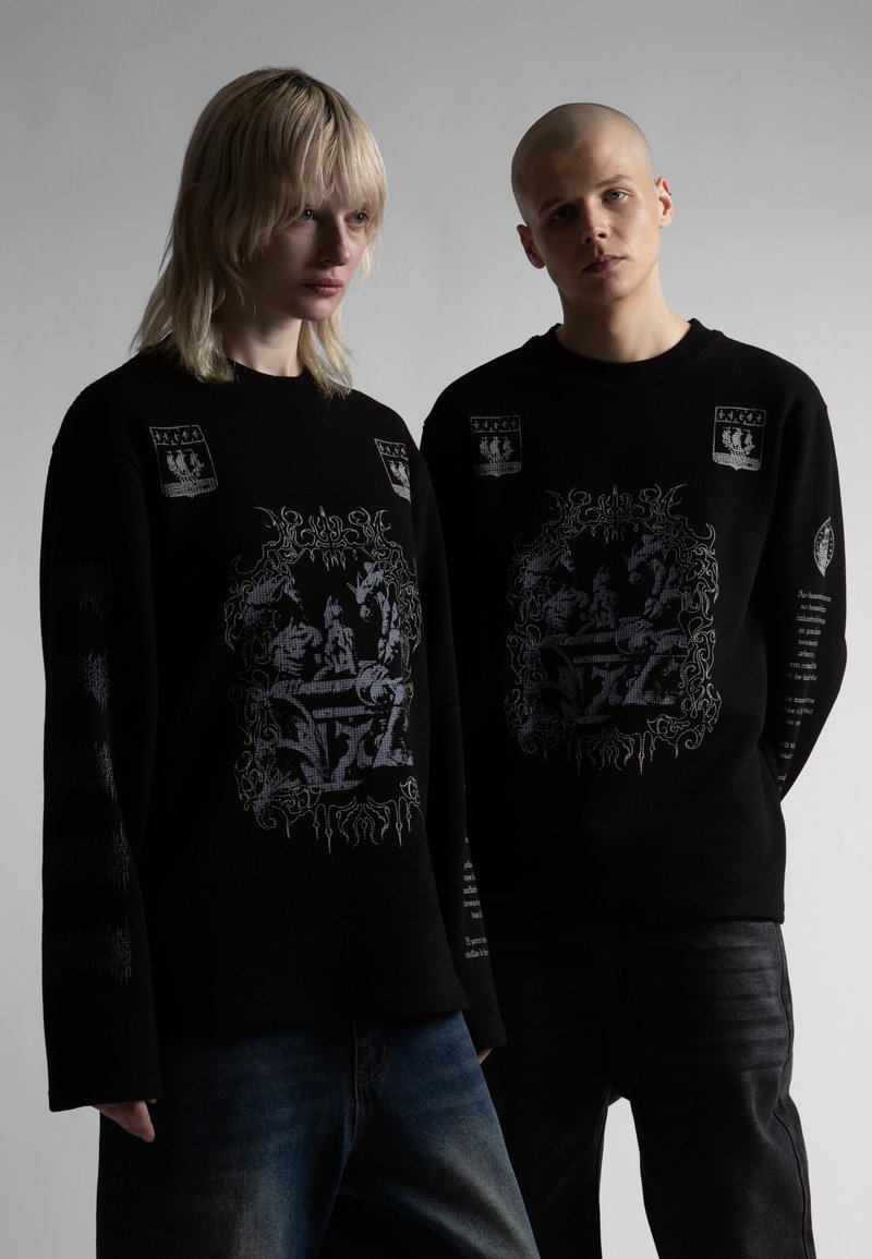 Δύο νεαροί ενήλικες φορούν μαύρα φούτερ με περίτεχνα μεσαιωνικού στιλ γραφικά, στέκονται μπροστά σε μια απλή γκρι φόντο.