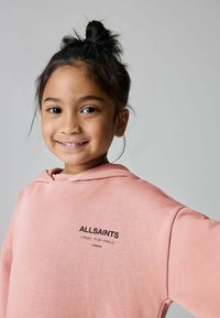 Roza dukserica s kapuljačom izrađena od teksturirane tkanine, s crnim logotipom koji na prednjoj strani kaže "ALLSAINTS". Jednostavan dizajn.