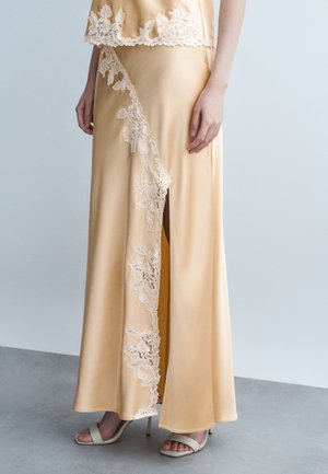 Beżowa satynowa spódnica z koronkową kwiatową lamówką wzdłuż przedniego rozcięcia, zestawiona z dopasowanym topem i białymi sandałami na obcasie.