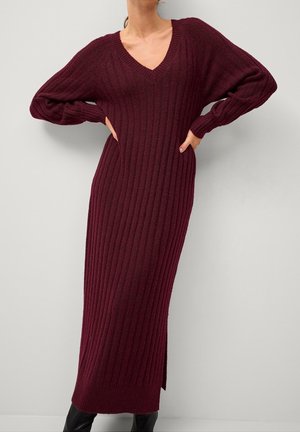 Nainen pukeutuneena pitkään, ribbattuun viininpunaiseen neulemekkoon, jossa on V-pääntie ja pitkät hihat, seisomassa käsivarret helmassa vaaleanharmaan taustan edessä.
