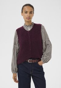 Bordeaux gebreide vest met V-hals en knopen aan de voorkant, gelaagd over een zwart-wit geblokt blouse met gerimpelde kraag en puffmouwen.