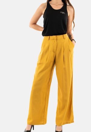 Woman wearing mustard yellow wide-leg pants, black sleeveless top, and black high-heeled shoes, standing with hands in pockets.