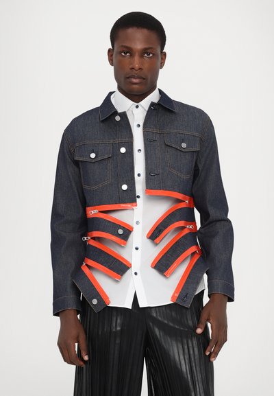 Veste en denim au design court, avec des accents de fermeture éclair orange et des détails de boutons. Assortie avec une chemise blanche et un pantalon plissé noir.