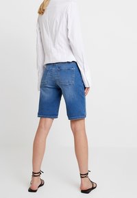 Denim bermuda shorts in medium blue, featuring a relaxed fit, slightly frayed hem, and back pockets. Paired with a white long-sleeve shirt.