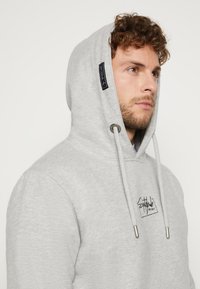 Grijze hoodie van zacht materiaal, met een trekkoordhoed, zilveren hardware en een geborduurd logo op de voorkant.