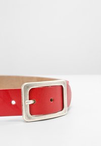 Sarkans ādas josta ar platumu, kurai ir sudraba metāla sprādze un vairāki caurumi izmēra regulēšanai. Plakanā tekstūra un pulēta virsma.