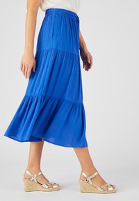 Falda azul, hasta la rodilla, con un diseño en capas. Hecha de una tela ligera, con una cinturilla elástica y una textura fluida. Combinada con sandalias de cuña doradas.