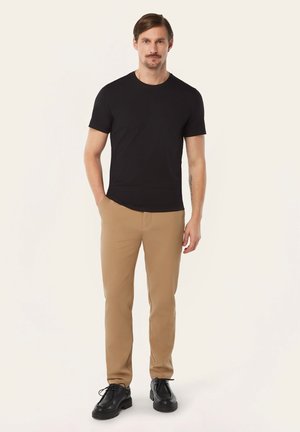 Man staat en draagt een zwart T-shirt met korte mouwen, beige broek en zwarte veterschoenen, met één hand in de zak tegen een effen achtergrond.
