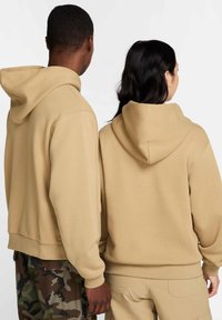 To personer har på seg beige hettegensere med løs passform, ribbede mansjetter og uten synlige logoer. Den ene har kamuflasjebukser, den andre har bukser i tan-farge.