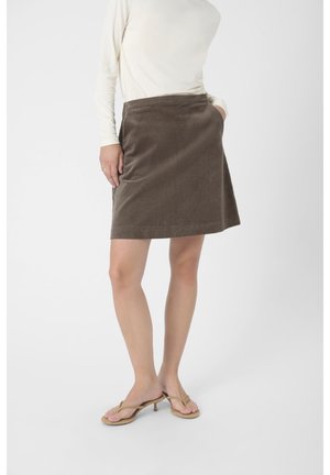Jupe en velours côtelé marron avec une taille cintrée, atteignant le milieu de la cuisse. Elle possède deux poches latérales et une texture lisse, accompagnée de sandales beiges.